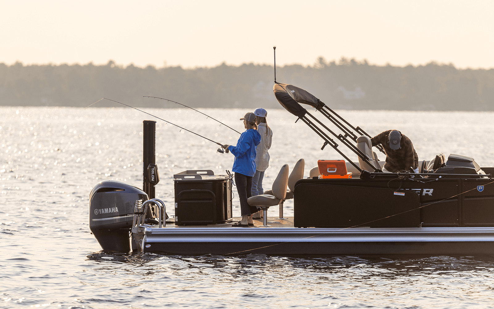 Two anglers casting directly over the water from the open aft deck of a Premier pontoon at sunset, with a Yamaha outboard at the stern